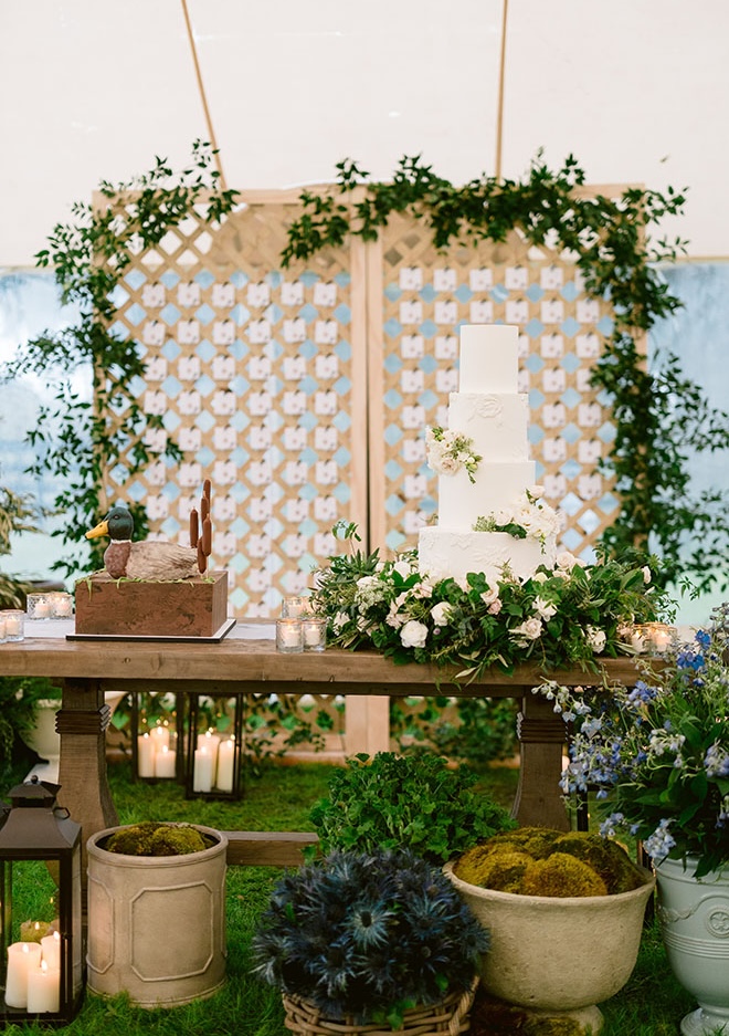 The groom's cake and wedding cake sit on a table in front of the seating chart surrounded by greenery.