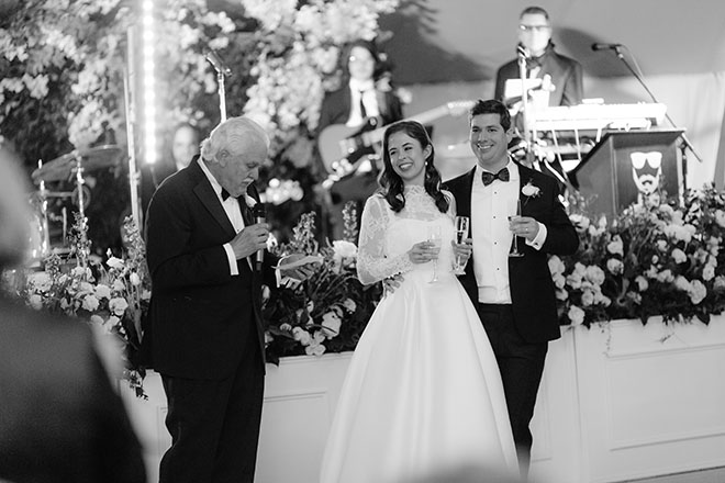 The bride and groom smile as the father of the bride shares a speech at the wedding reception. 