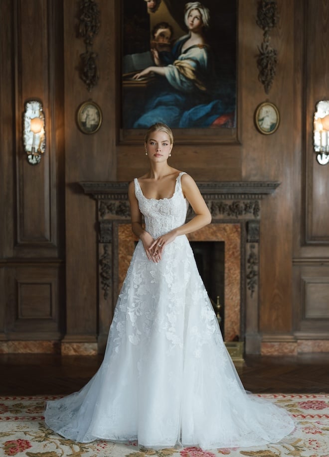 A bride wearing the Anne Barge Oakland gown features a scoop neck A-line silhouette with a low back adorned with intricate floral embroidery and a cathedral length veil with vine-embroidery on the trim.