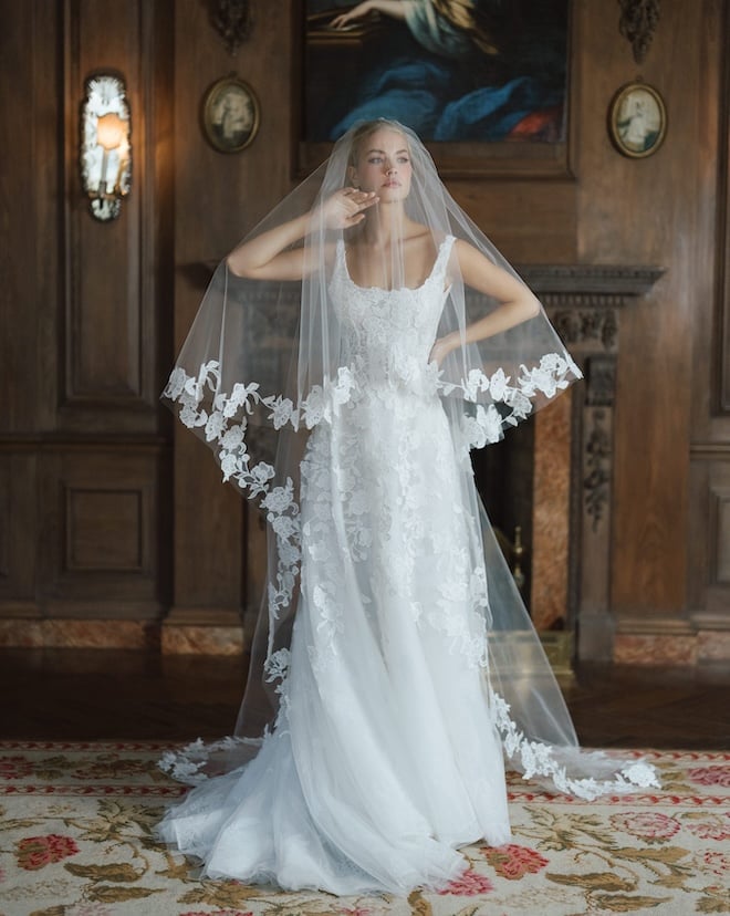 A bride wearing the Anne Barge Oakland gown features a scoop neck A-line silhouette with a low back adorned with intricate floral embroidery and a cathedral length veil with vine-embroidery on the trim.