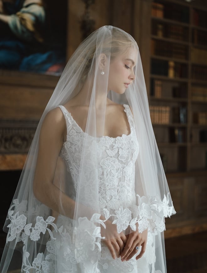 A bride wearing the Anne Barge Oakland gown features a scoop neck A-line silhouette with a low back adorned with intricate floral embroidery and a cathedral length veil with vine-embroidery on the trim.