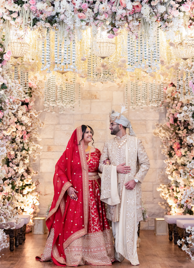 The bride and groom smiling at each other under the floral Mandap. 