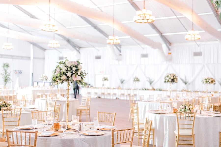 A pavilion venue adorned in white decorating reception tables, white florals, and bright ceiling lighting. 