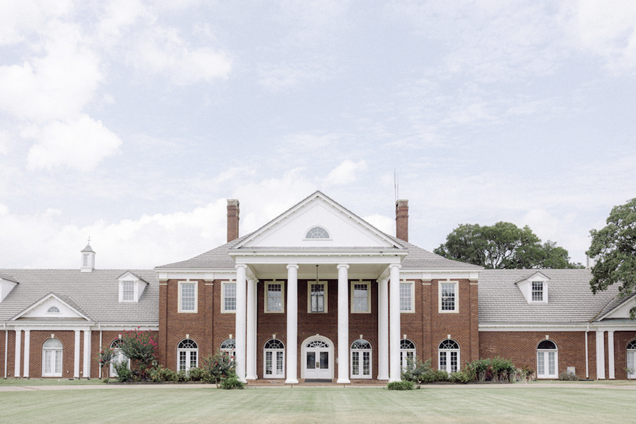 A mansion stands tall in Texas Hill Country with tall white pillars and lush greenery. 