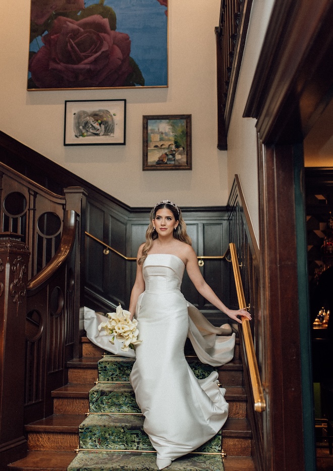 The bride walks down the stairs wearing her wedding gown and holding her bouquet.