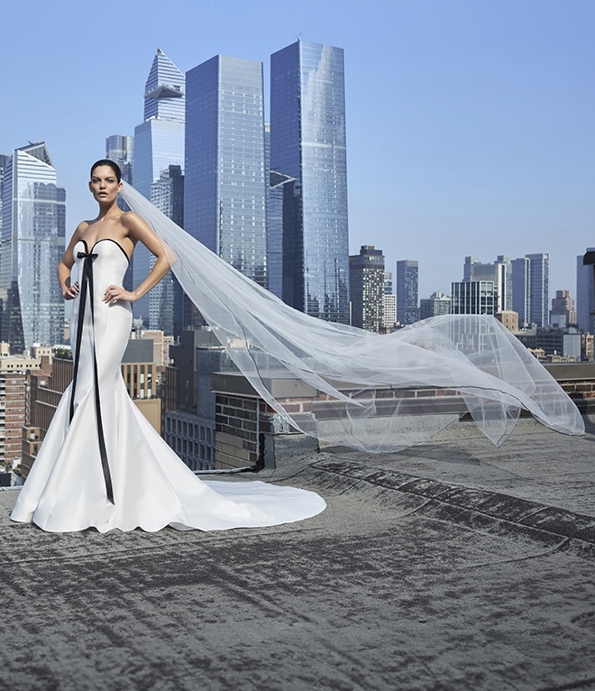 The model stands on a rooftop overlooking the city wearing a white wedding gown with a black bow. 