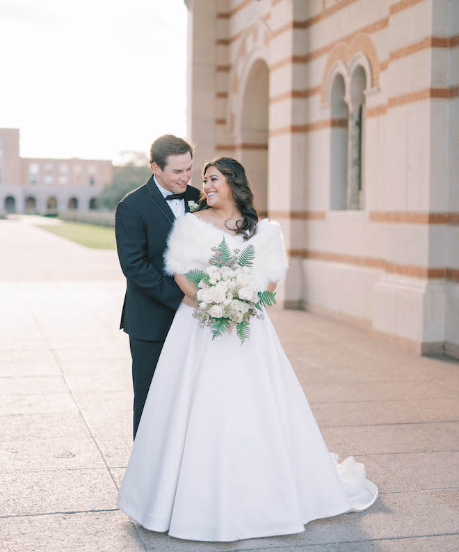 The bride and groom smile while taking their wedding portrait photos in front of a church. 