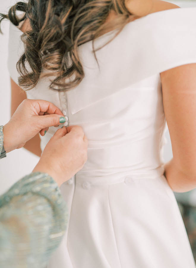 A friend helps the bride button the back of her dress before she gets married in her satin white wedding gown. 