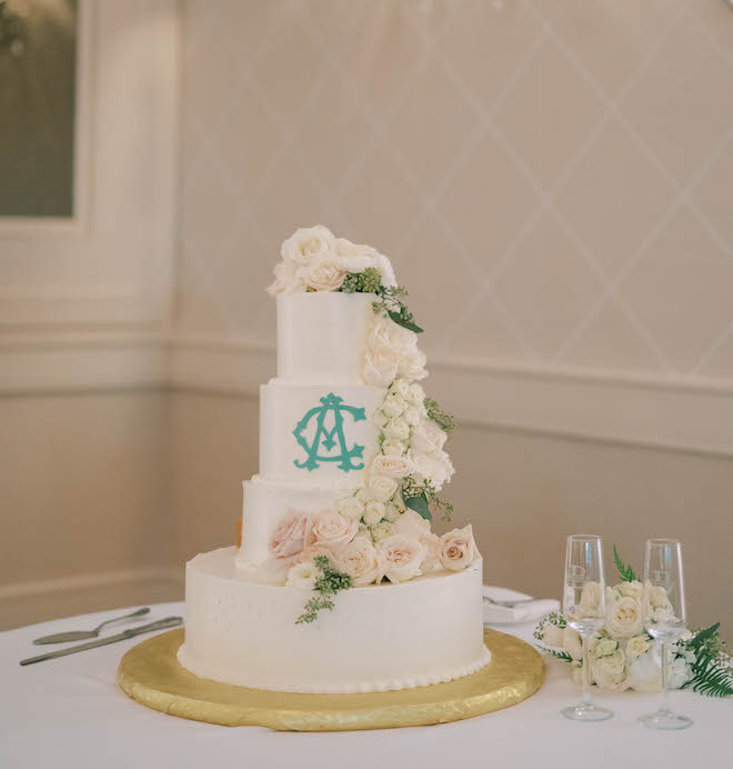 A white four-tier wedding cake with light blush pink and white florals align the side. Featuring light blue monogrammed letters. 