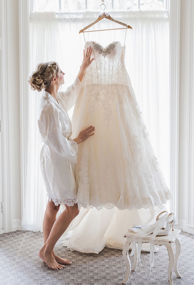 The bride looks up at her wedding dress that hangs in front of a window. 