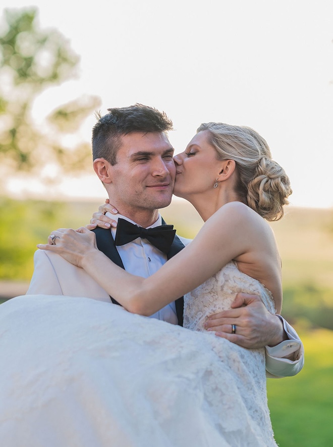 The bride gives her groom a kiss on the cheek as he carries her around outside. 