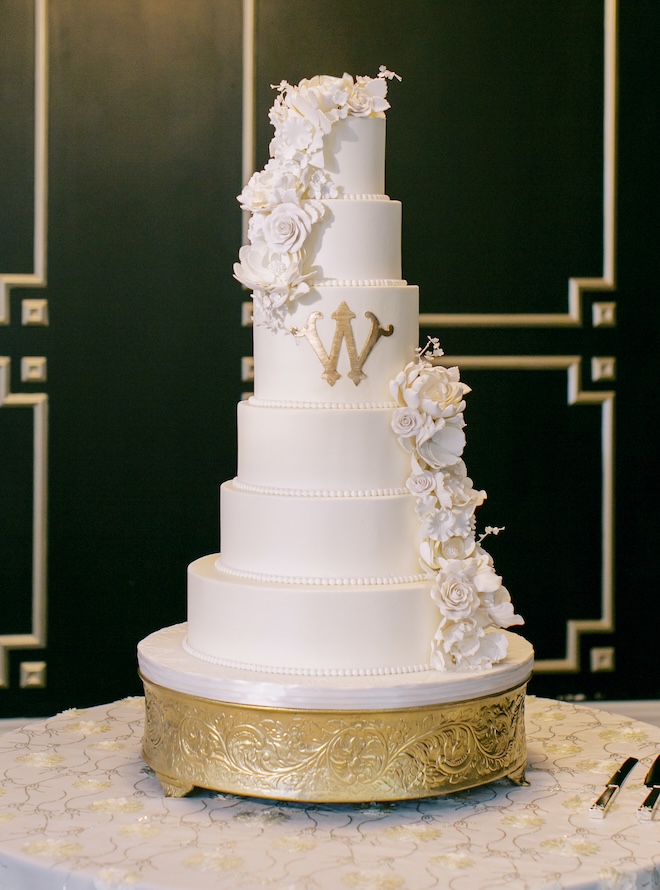 Cakes by Gina designs a six-tier white  custom wedding cake with white flowers and gold detailing. 