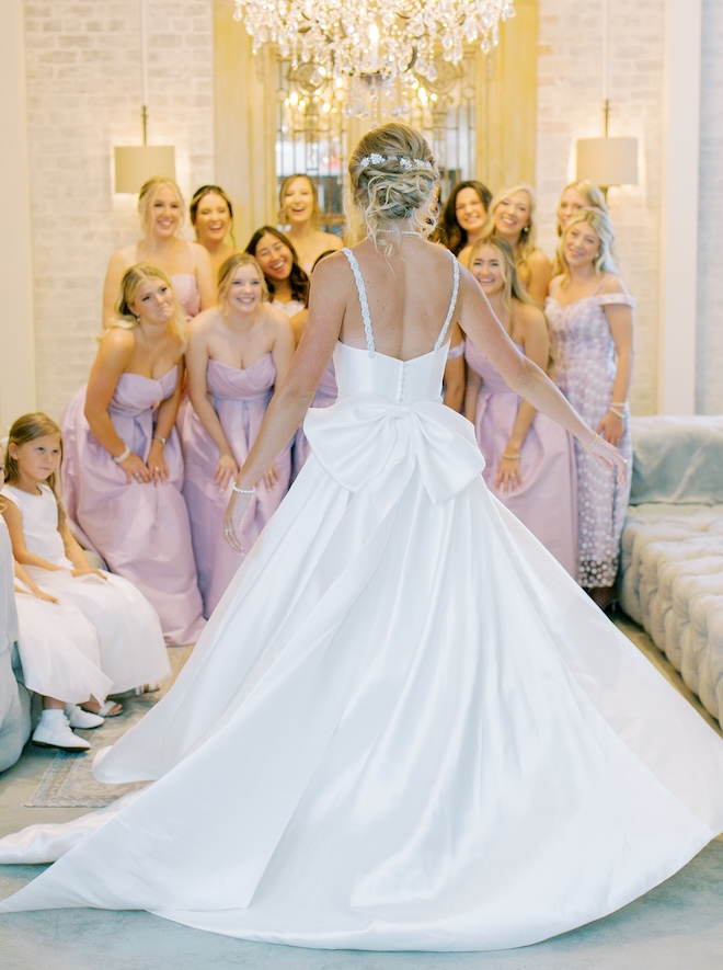 The bride wearing a sleeveless gown with a bow in the back revealing her dress to her bridesmaids. 