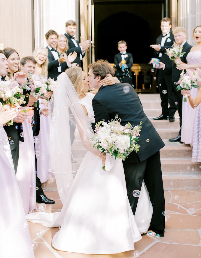 The bride and groom kissing outsidr the church with guests cheering and blowing bubbles. 