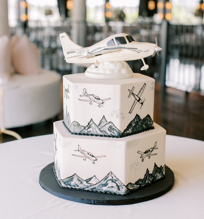 A two-tier groom's cake with planes flying over mountains and a plane replica on top. 