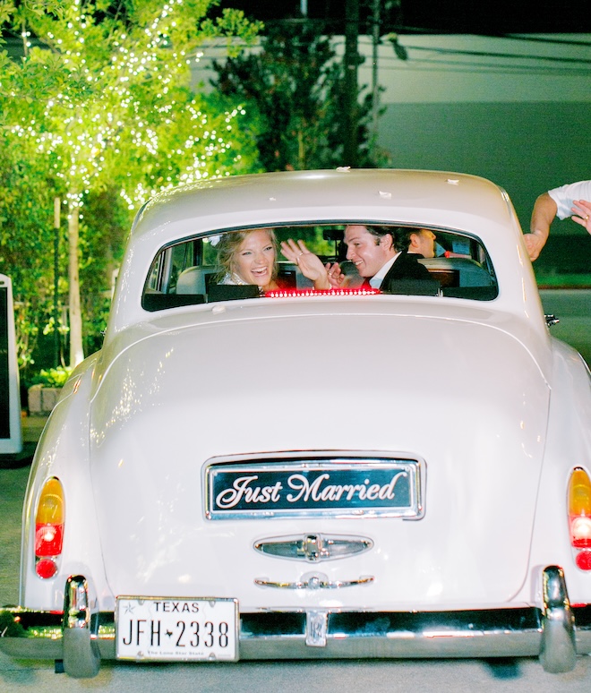 The bride and groom waving from the back window of a white vintage car with a license plate that says "Just Married". 