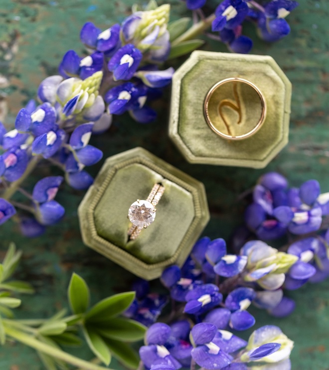 An engagement ring on a green velvet ring box next to bluebonnets. 