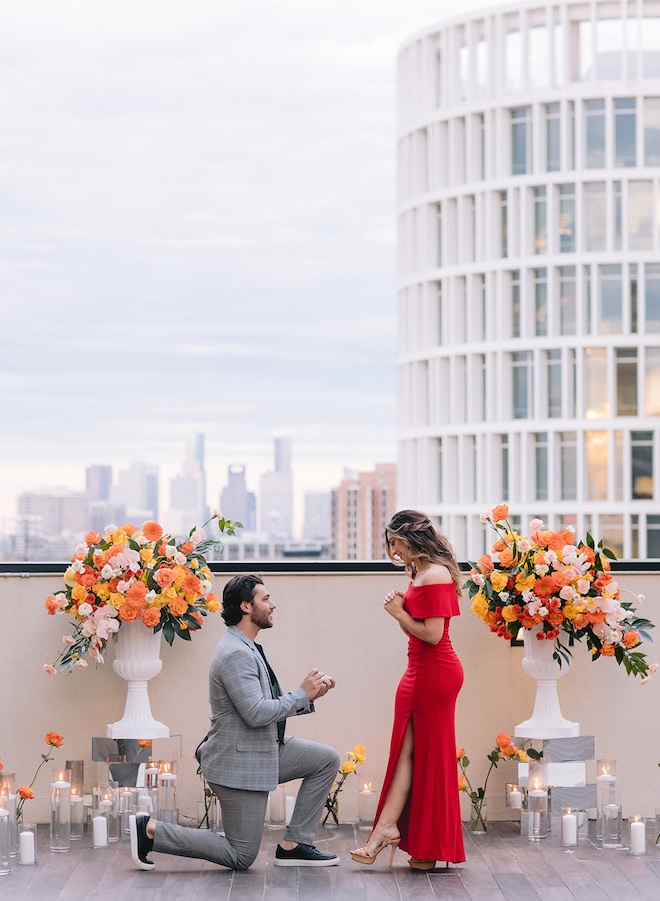 A man on one knee proposing to a girl on the rooftop overlooking the skyline.