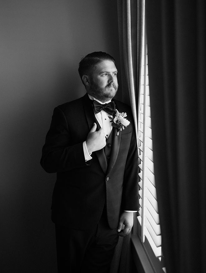 The groom looks out the window while wearing a tuxedo.