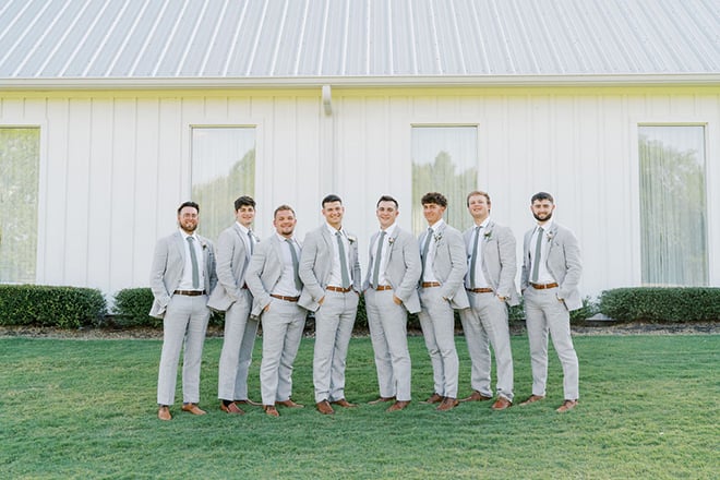 The groom and his groomsmen stand outside the white farmhouse wedding venue. 