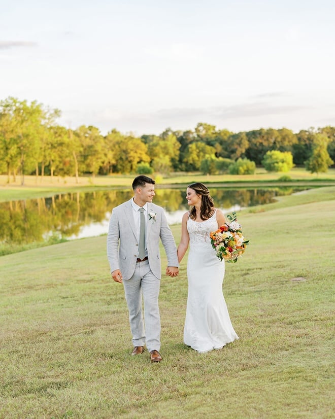 The bride and groom hold hands in front of a pond in an open field. 