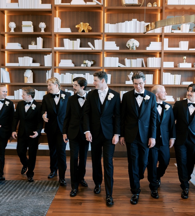 The groomsmen walking in front of brown bookshelves with white books. 