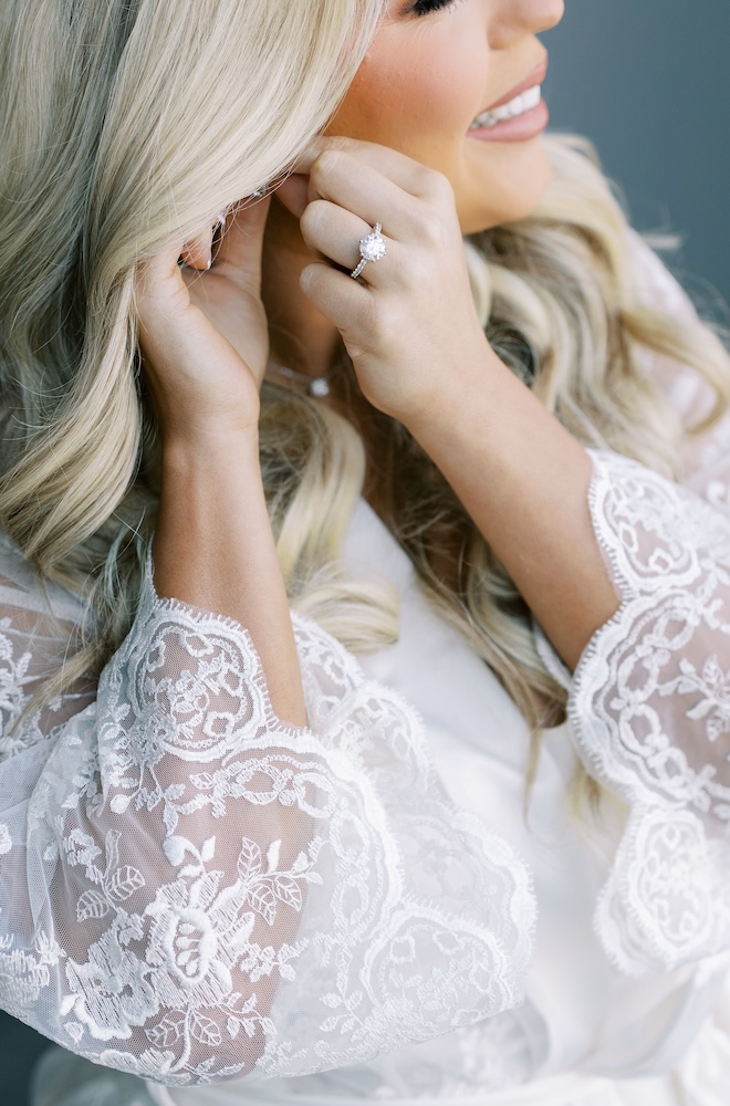 The bride wearing a white lace robe putting on her earrings. 