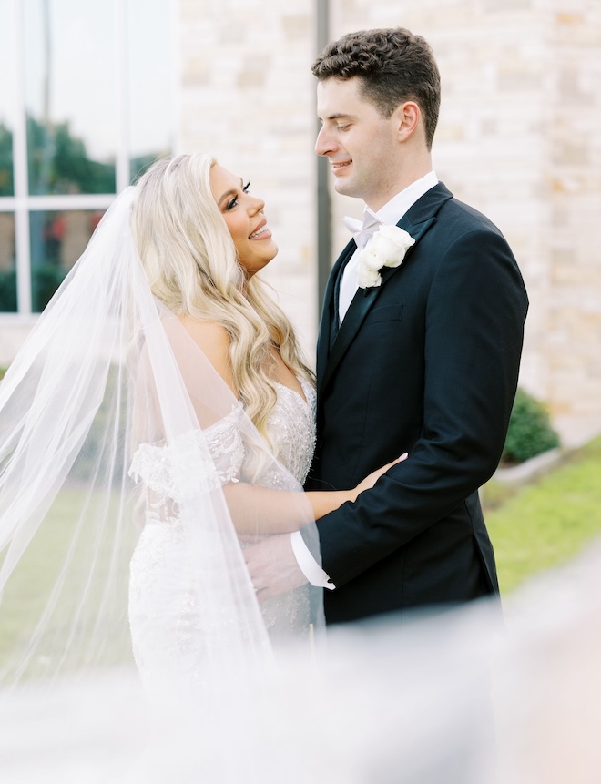 A bride and groom looking at each other smiling outside a church. 