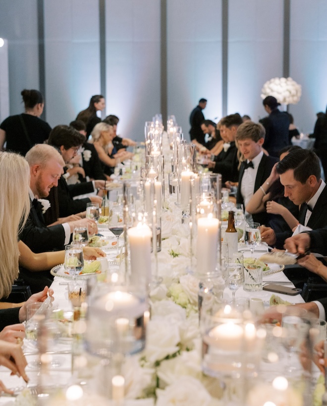 Guests dining at a long reception table lit with candles and white florals. 