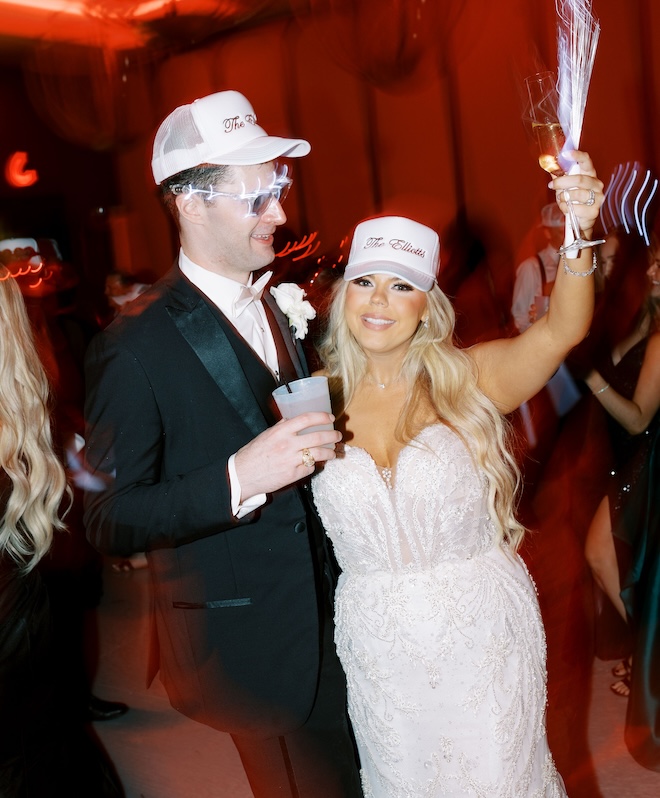 The bride and groom wearing hats and holding up their drinks during their wedding reception.