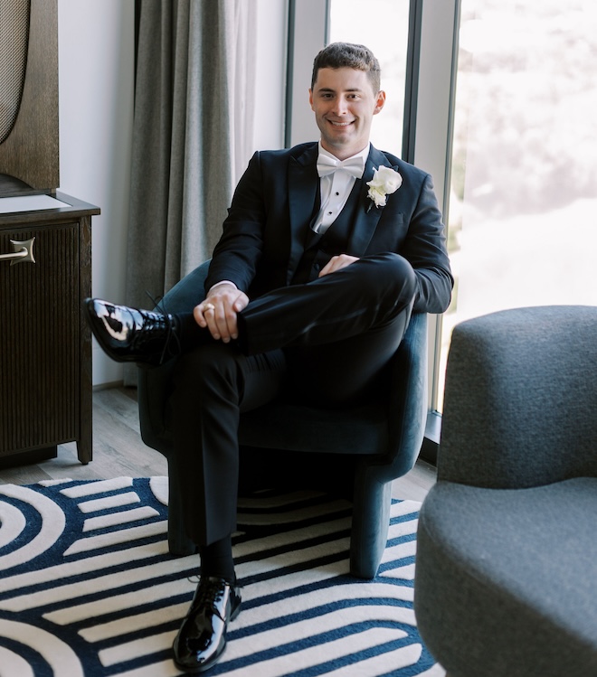 The groom smiling in his tux while sitting in a room at Thompson Houston, by Hyatt. 