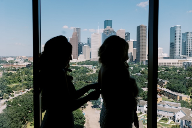 The silhouette of a bridesmaid zipping the brides dress with the Houston skyline in the background. 