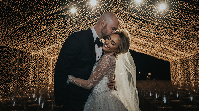 The groom Ryan Pressly kissing his brides cheek under a canopy of lights. 