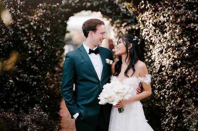 The bride and groom smiling at each other underneath a grand hedge archway. 