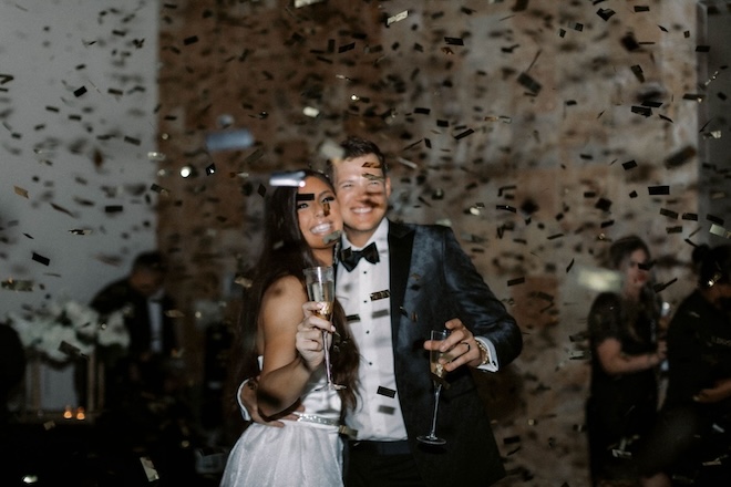 A bride and groom smiling while holding champagne with confetti raining on them. 