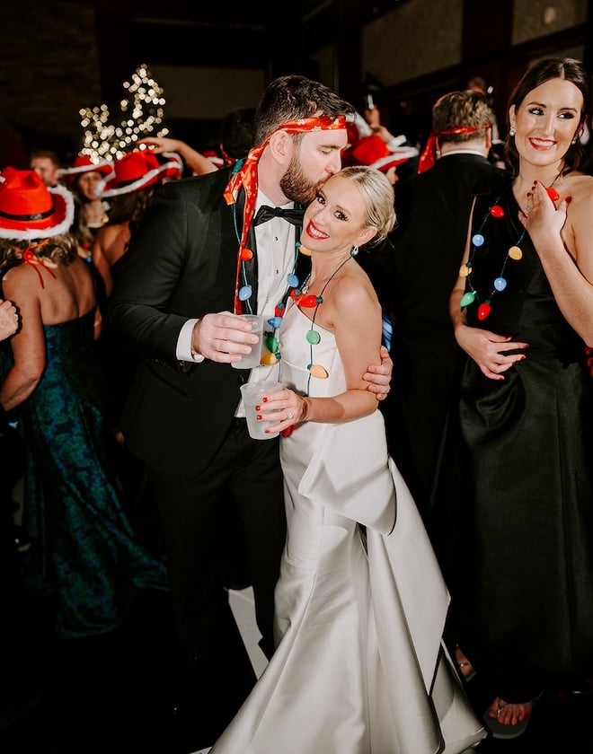 The groom wearing his tie as a headband kissing the bride wearing a Christmas light necklace. 