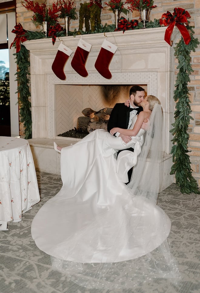 The bride and groom kissing at the fireplace with three stocking hung above them. 