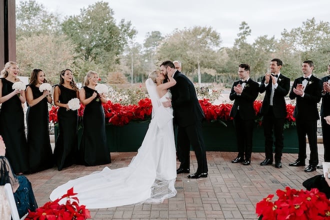 The bride and groom kissing at the altar while the wedding party claps behind them. 