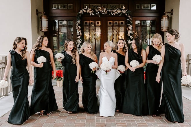 The bride and bridesmaids wearing black walking in front of a Christmas decorated door.