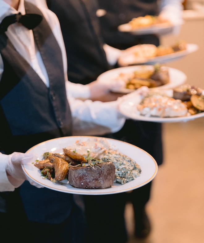 Cater waiters holding plates of food including, steak, potatoes and creamed spinach. 