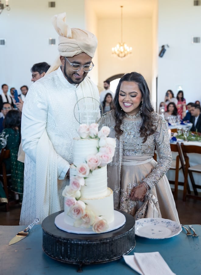 The bride and groom cutting into a 3-tier wedding cake with pink roses on it. 