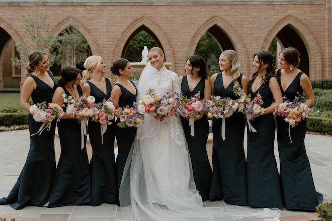 The bride smiling with her bridesmaids wearing black dresses with colorful bouquets. 