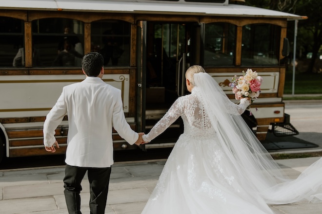 The bride and groom holding hands walking toward a brown and white trolley. 