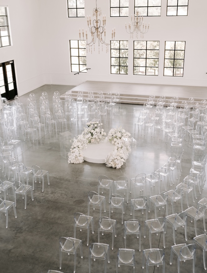 The ceremony space with white florals surrounded by white clear chairs. 