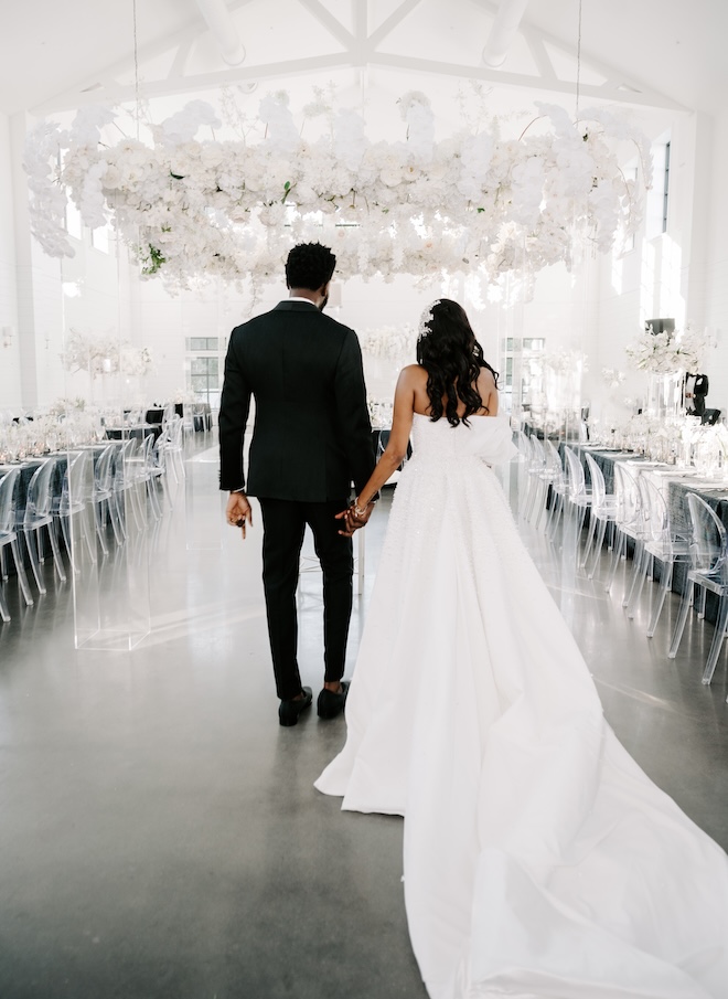 The bride and groom walking into their reception space underneath a white floral arrangement hanging above them. 