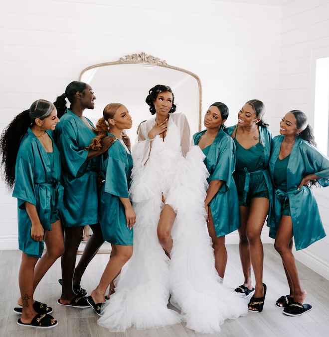 The bride in a white robe posing with her bridesmaids in teal robes.