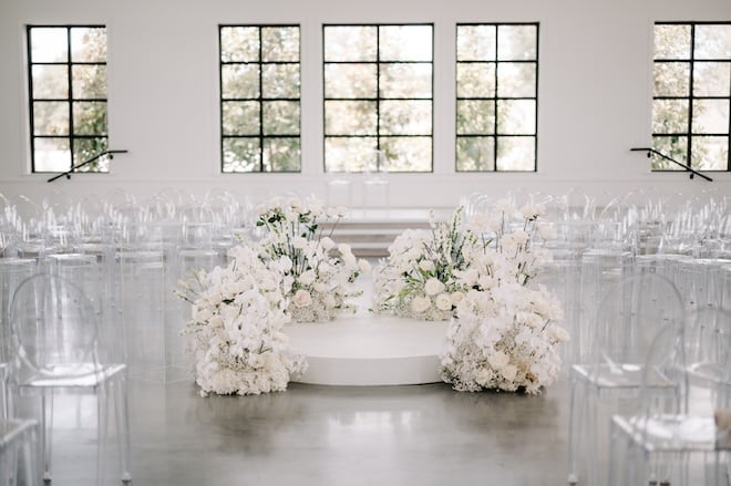 The ceremony space with white florals surrounded by white clear chairs. 