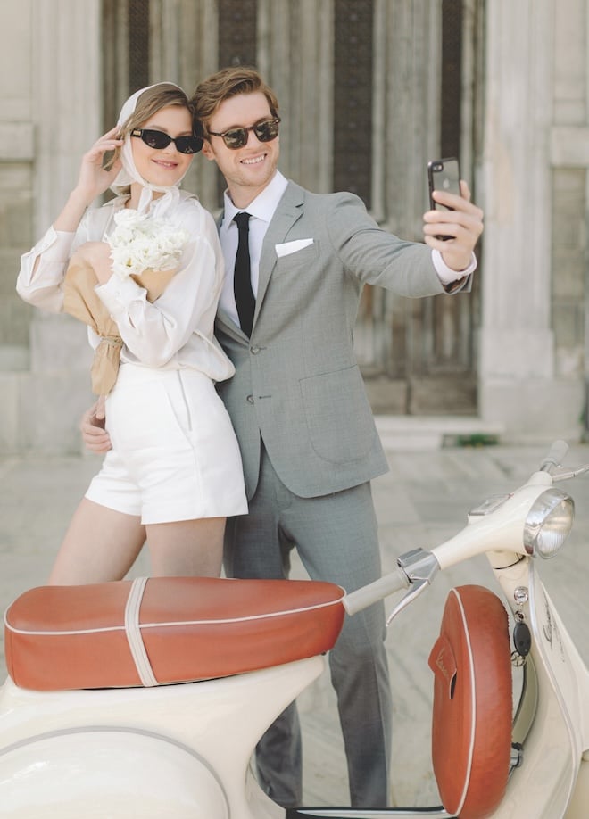 A couple smiling taking a selfie with a cream vespa in front of them. 