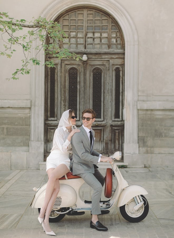 A couple smiling while posing on a cream vespa. 