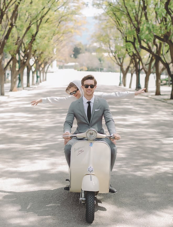 A couple writing a cream vespa along a road lined by trees. 